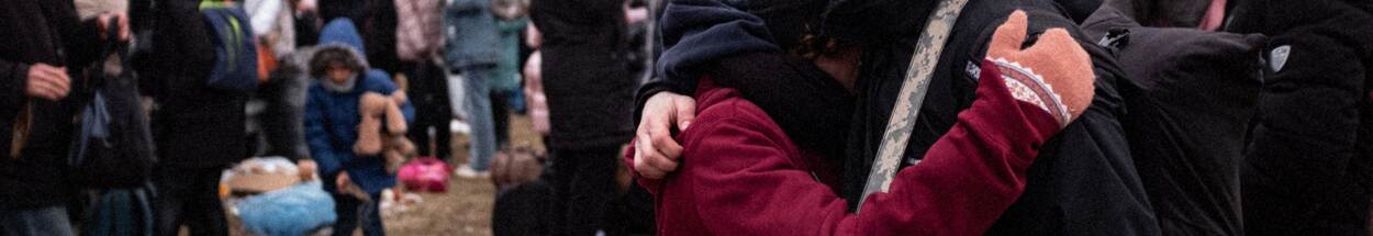 2 vluchtelingen omarmen elkaar met op de achtergrond andere vluchtelingen, waaronder een kind met een knuffel