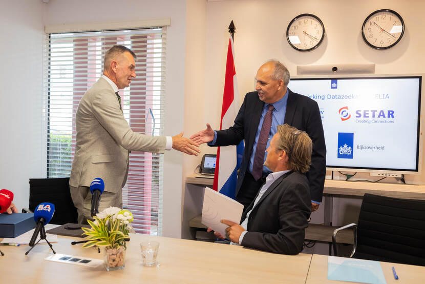 Staatssecretaris Eddie van Marum (links) geeft SETAR directeur Roland Croes een hand tijdens het persmoment op Aruba, terwijl SSCS directeur Werner (zittend) Haan toekijkt.