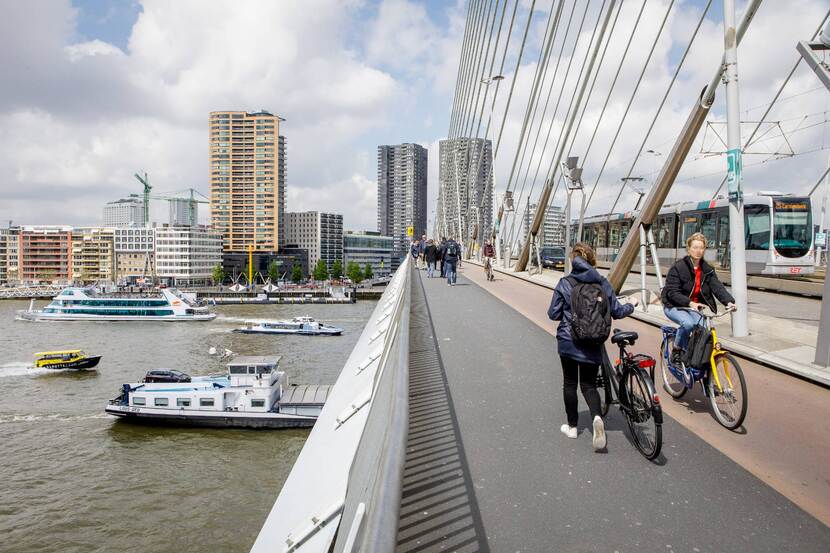 Voetgangers, fietsers en een tram op de Erasmusbrug. Onder de brug varen enkele schepen.