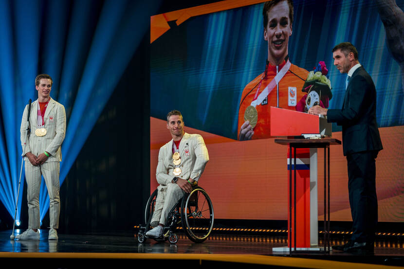 Op een podium staan twee sporters met medailles, een van hen zit in een rolstoel. Iemand anders geeft een toespraak achter een katheder.