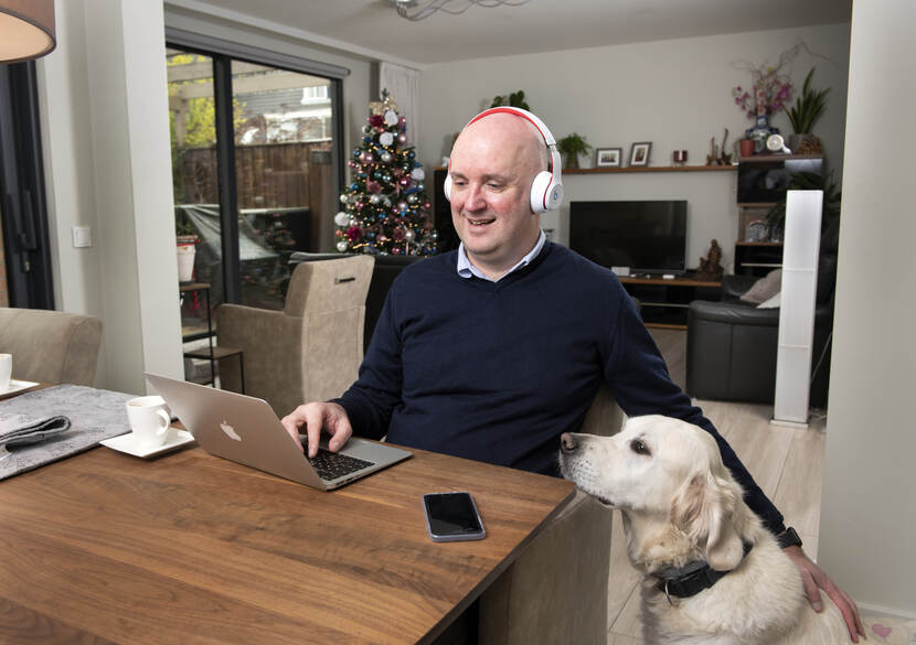 Een foto van een man achter een laptop die zijn hond aait. Op de achtergrond staat een kerstboom.