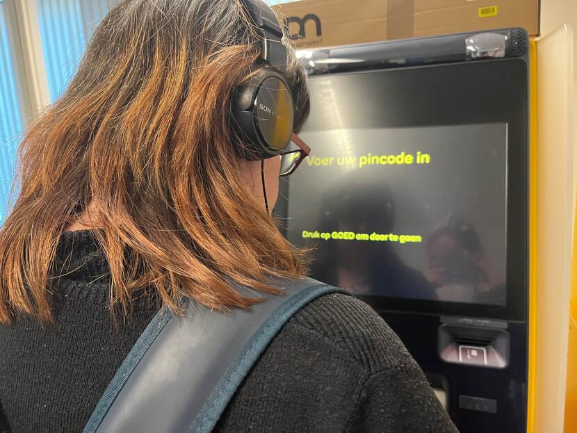 Vrouw met visuele beperking met koptelefoon op staat bij een geldautomaat met grote letters en veel contrast